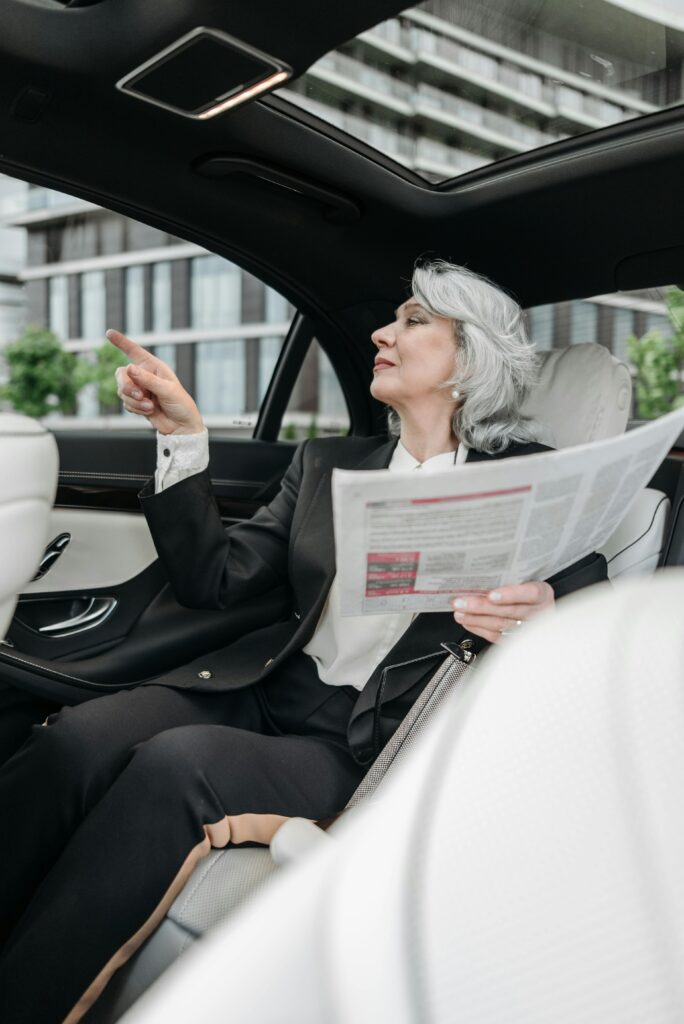 Elegant senior businesswoman points while reading a newspaper in a modern car interior.